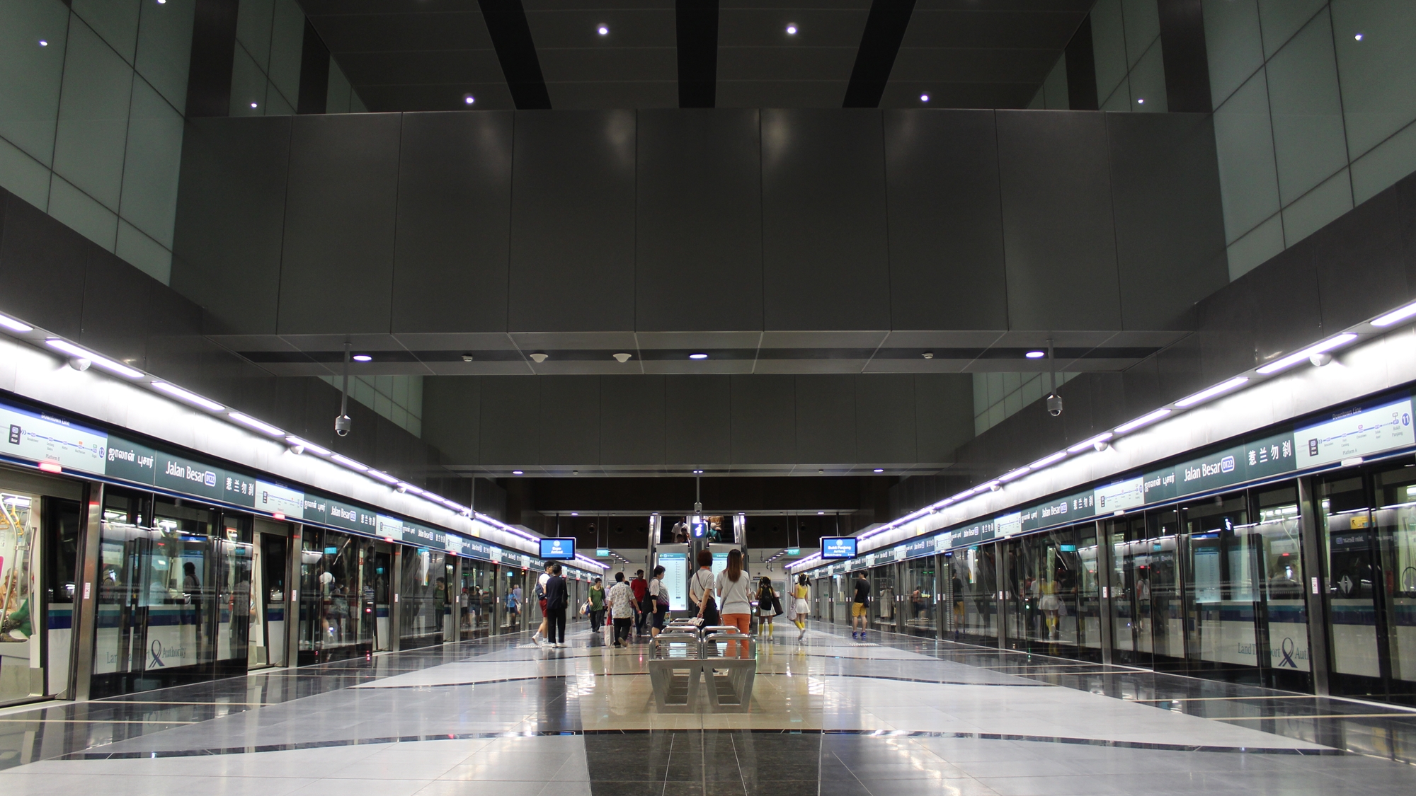 A symmetrical view of the MRT platform at Jalan Besar featuring dark, metallic grey ceiling panels and bright white floor tiles. The platform is relatively quiet with a few distant figures.