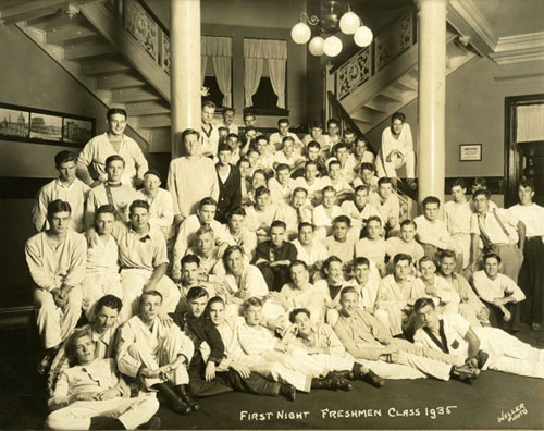 About 50 members of the 1935 freshman class gather on the first floor of their dormitory, which has columns, a chandelier with spherical lights, and multiple staircases.