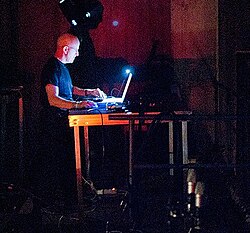 slightly pixellated, distant image of Brian Williams standing at DJ station from right profile, wearing a dark t-shirt