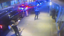 A dirty frame of CCTV footage covering the sidewalk. The killer stands at the bottom left, wearing a hooded jacket, firing his sidearm at Thompson, in the center, his back facing the camera.