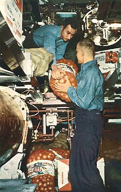 U.S. Navy enlisted man lifting a bag of potatoes surrounded with other bag and cases of food in a submarine torpedo room.