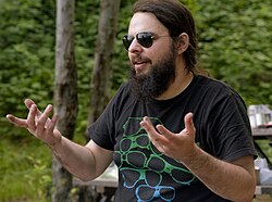 McGrath gesticulates with both hands while looking to the left. He wears aviator sunglasses, a black T-shirt with a sunglasses graphic, and has a long, brown beard. He is in an outdoors, camping setting.