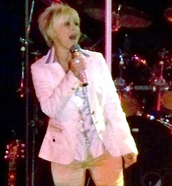 A blonde woman wearing a white jacket singing into a microphone