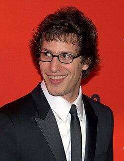 An upper body photo of Andy Samberg in 2010. He is wearing a black suit, white shirt, and black tie. He is smiling wide and looking to the left of the photographer.