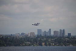 Six fighter planes fly in a tight triangle formation above city skyline on a cloudy day