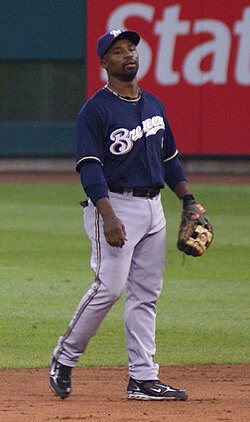 A man wearing gray pants, a navy blue jersey with "Brewers" written in white, a navy blue cap bearing a white "M", and a baseball glove stands on a baseball field.