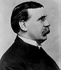 A mustached man wearing a dark suit and pince-nez glasses looks to the right