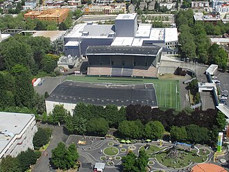 Aerial view of a stadium with two covered sections sited between a playground and other buildings in an urban neighborhood.