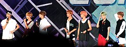 Seven men standing on a stage with each holding a microphone and dressed in casual, colorful outfits, with hairstyles varying from natural to brightly dyed colors like pink and green.