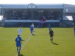 A football match with players and a referee on the pitch. A small spectator stand full of people is in the background.