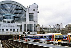 An NSE-branded Class 465, a commuter train, at a very grand terminus station. The skyline of London is visible in the background and about two-thirds of the station building. The photo is taken from quite a few platforms away