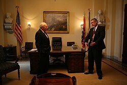  Cheney and Addington stand in front of the Wilson desk in a room with cream colored walls. A painting, and two busts hand on the wall.