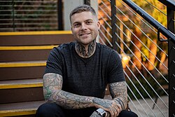 A smiling man in a black T-shirt sitting on some stairs; he is covered in tatoos