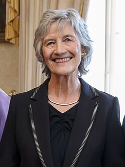 An above shoulder portrait of Catherine Connolly. She is a woman with grey and curly shoulder length hair. She is wearing a dark pinstriped blazer over a white blouse. She is smiling staring directly at the camera.