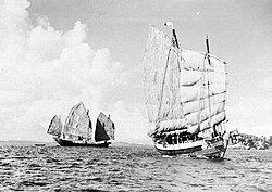 Black and white view of the ocean with an island visible on the horizon to the right. A sailing ship on the left (three sails visible) shows the full length of its hull while another on the right (two sails visible) shows its forward bow.