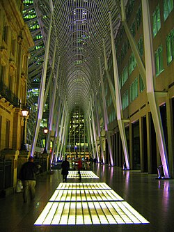 A towering metal gridwork catenary arch forms a gallery over a shiny concrete concourse inset with glowing grids of glass. Beyond the arch, city lights of tall buildings are visible. A four-story historic stone building stands to the left; a plain modern building rises out of sight to the right.