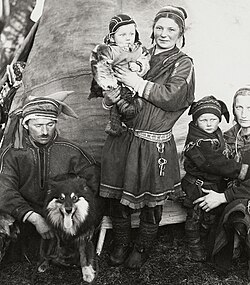 A black-and-white photograph of a Sámi family in Finland wearing traditional clothing, with adults and children gathered outside beside a dog, taken before 1936.