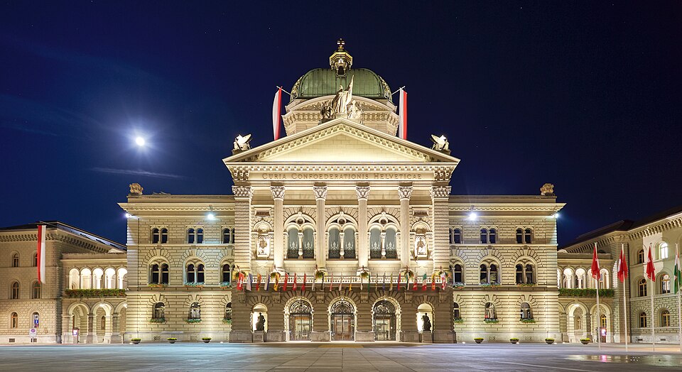 Federal Palace of Switzerland (Bundeshaus) – Pediment with inscription Curia Confoederationis Helveticae (assembly of the Swiss federation) – view including the allegorical sculpture of executive, independence, and legislative as well as the central dome crowned by the swiss cross – pictured during full moon with clear night sky.