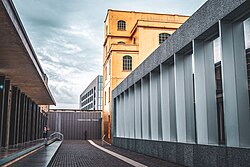 Under an overcast sky, a brick aisle runs between a brown building on the left, and a golden structure and array of glass windows split by grey columns on the right; in the near distance is a brown wall with white letters spelling out "FONDAZIONE PRADA". Another building, several stories tall, lies in the background.