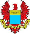 escudo azul cielo, no es el azul heráldico. Alas amarillas sobre fondo amarillo; pico cerrado y rojo igual que las uñas; letras negras.