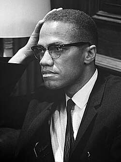 An African-American man in his forties, wearing glasses and a suit and tie, sitting and looking to the right, with his hand resting on his right temple.