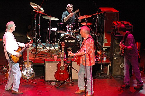 Three men playing guitars and one playing drums, performing together on a stage