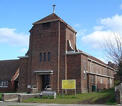 A red brick church stands with the small steeple facing the street as the church's entrance. The building extends back from the entrance, and the building is surrounded by grass.
