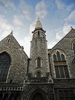 A picture of a church, built out of stone, with stained glass windows, viewed upwards from the street. Behind the church, a blue sky is lightly filled with white clouds.