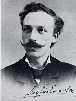 white man in 1900s costume, seated; he has wavy dark hair of medium length and substantial mustache