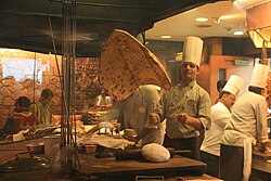 A chef in a chef uniform holding a very large naan using two rods.
