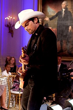 Brad Paisley, dressed in a black suit and white cowboy hat, and playing an electric guitar in a formal setting