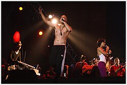 A shirtless man in the front holding a microphone, another man with a black vest in the right playing the keyboards, a woman in the left holding a microphone, and a musical ensemble playing several instruments in the back.