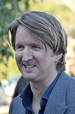 A man in a grey suit with a blue shirt looks down to his right while smiling.