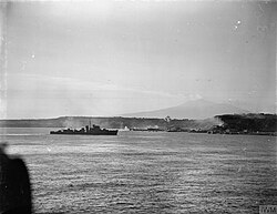 A black and white photograph showing a warship firing its armament at positions on the foreshore, while in the distance landing craft move towards the beach