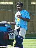 A man in a blue t-shirt, wearing white tracksuit trousers with pads. A grass-cutting machine can be seen in background.