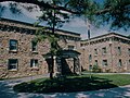 A flagpole and pine tree in front of a large two story stone building with many windows