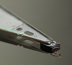 Close-up of an HDD head resting on a disk platter; its mirror reflection is visible on the platter surface. Unless the head is on a landing zone, the heads touching the platters while in operation can be catastrophic.
