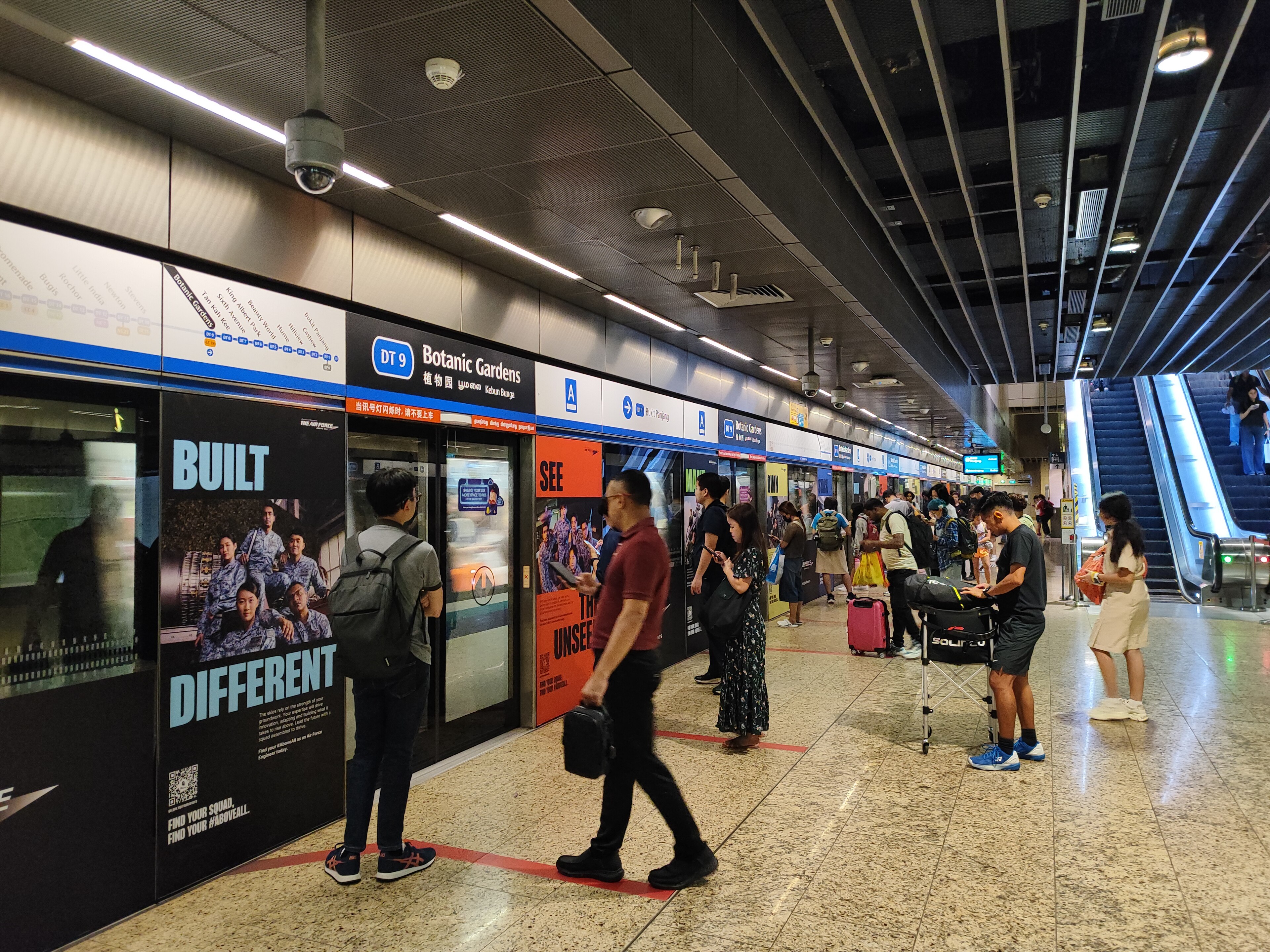 Commuters wait for the incoming train on a brightly lit platform at Botanic Gardens featuring glass screen doors and visible escalator access.