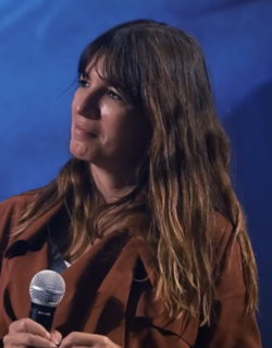close-up of Lúcia Moniz in left semi-profile, wearing a dark brown shirt, holding a microphone, and smiling with head slightly tilted to the left