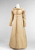 A headless mannequin wearing a light beige floor-length coat that buttons up the middle with three lines of decorative, zigzagging cording along the fitted bodice, long sleeves, and scalloped collar