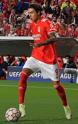 A man with a red shirt and white shorts running with a football