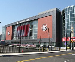 A red and black building with large glass towers on either side is seen in the background. The words "Prudential Center" are on top in white. A parking lot and street are visible in the foreground.
