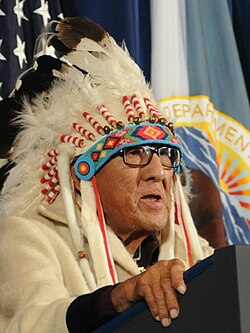 Joe Medicine Crow in full feathered headdress speaking while putting his hands on a podium