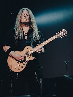 Roland Grapow wearing dark pants and a dark, open-necked shirt, standing onstage, playing electric guitar with eyes closed and pursed lips