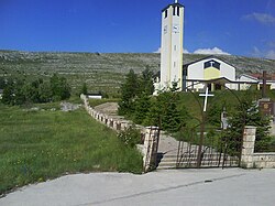 A photo of a white church on the outskirts of a village, taken from outside the front gate of the church, just outside of its garden. In the background is a stony hill.