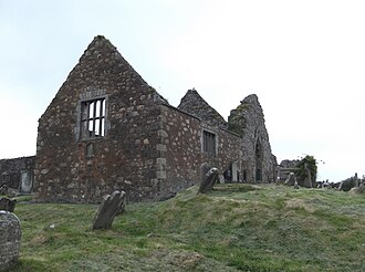 A photo of a ruined stone building with a gable and a mullioned window standing in a graveyard