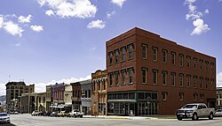 A group of older buildings line a downtown street, several vehicles are parallel parked in front of them and a few clouds can be seen in the sky