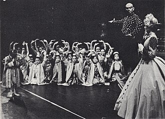 Black and white photo of a theatrical scene: a middle-aged man with a shaved head and imposing presence has his right arm extended to introduce a group of children in Asian dress to a woman in a crinoline dress and bonnet in the foreground at right, who is partially turned upstage.  The children are mostly kneeling and have their arms raised in greeting; one child (probably Crown Prince Chulalongkorn) stands and bows.