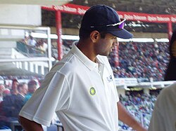 A male, dark-skinned cricketer in a white short-sleeved shirt and dark blue baseball cap with sunglasses on the peak of the cap. He is standing high up in a cricket stand with a large number of spectators in the background.