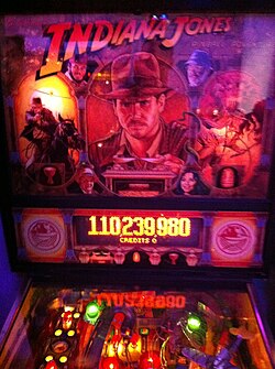 The image contains a backbox of a Indiana Jones themed pinball machine. The backbox shows a image of Indiana Jones and below it with a screen telling the player they got 110,239,980 points.